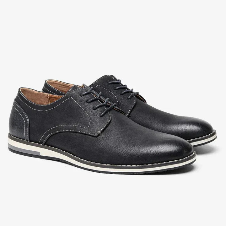 A pair of black leather patchwork oxfords with white stitching, lace-up fronts, and low white and black rubber soles, featuring a cushioned sole for added comfort, displayed on a light background.