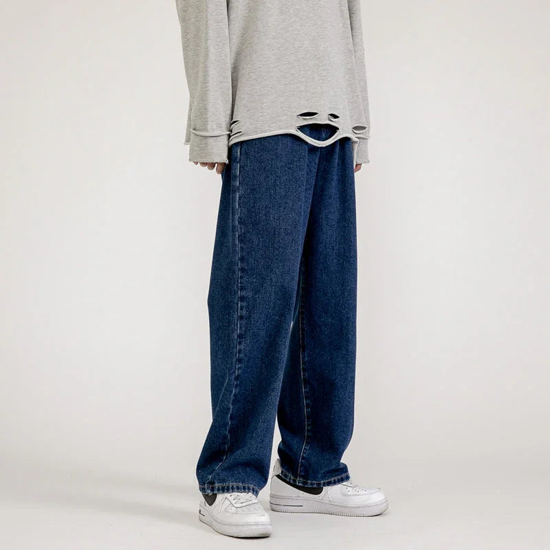A person wearing a loose, light gray long-sleeve shirt with a distressed hem and men’s baggy jeans in a wide-leg design, paired with white sneakers, channels effortless Korean fashion against a plain background.