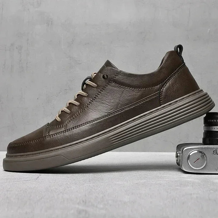 A brown genuine leather sneaker with beige laces and a textured rubber sole is displayed sideways on a gray surface, with part of a vintage camera visible in the foreground.