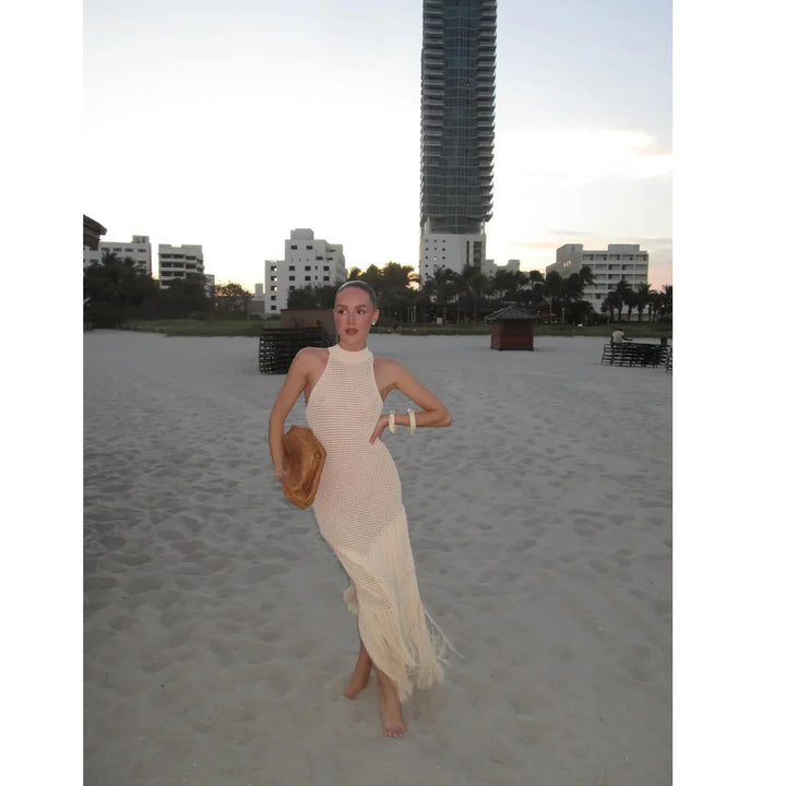 Woman in a white crochet dress on a sandy beach at sunset, city buildings in background, Rovelli
