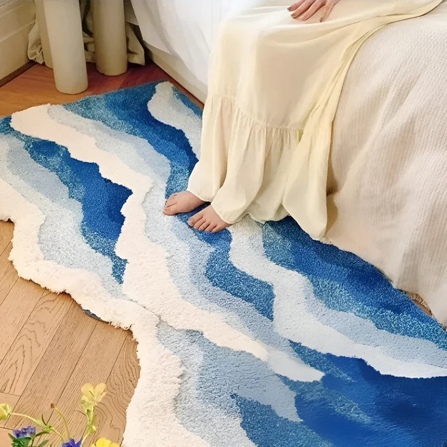 Woman in cream dress sits on bed, feet on wavy blue and white Rovelli rug on wood floor