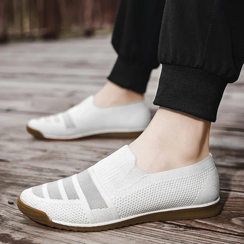 A person wearing lightweight slip-on mesh sneakers with brown soles stands on a wooden surface. The breathable knit shoes feature a cushioned insole, and the person is dressed in black cuffed pants.