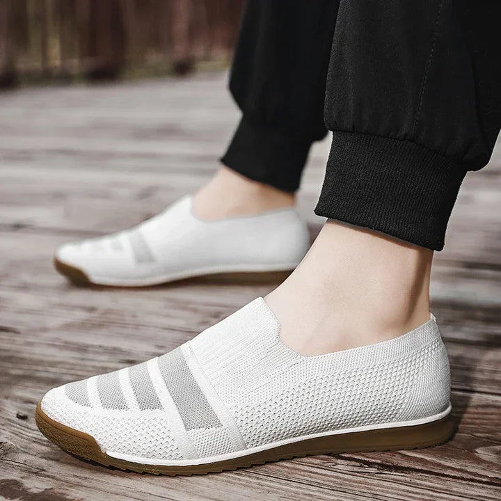 A person wearing lightweight slip-on mesh sneakers with brown soles stands on a wooden surface. The breathable knit shoes feature a cushioned insole, and the person is dressed in black cuffed pants.