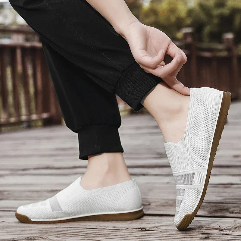 A person standing on a wooden deck outdoors wears black pants and white lightweight slip-on shoes with light brown soles, adjusting one shoe by the heel with their hand.