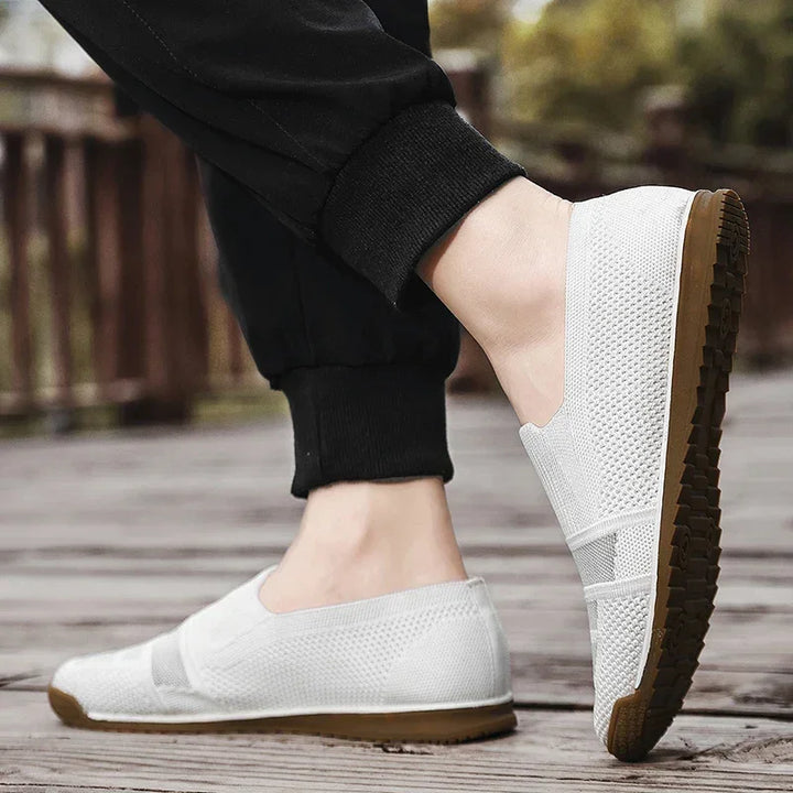 A person wearing lightweight slip-on mesh shoes with brown soles stands on a wooden surface outdoors. The black pants complement the shoes, while blurred greenery and a fence appear in the background.