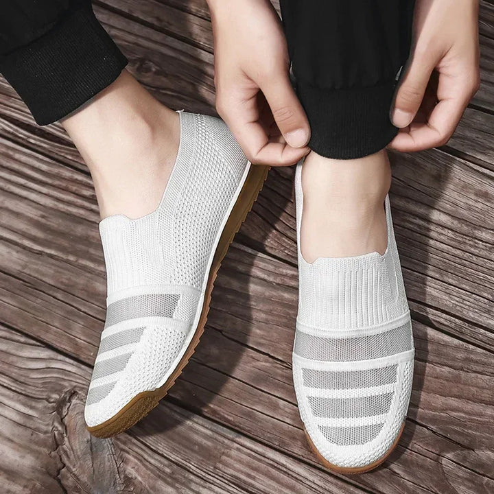 A person wearing lightweight slip-on knit sneakers with light brown soles is adjusting the cuff of their black pants while standing on a wooden surface.