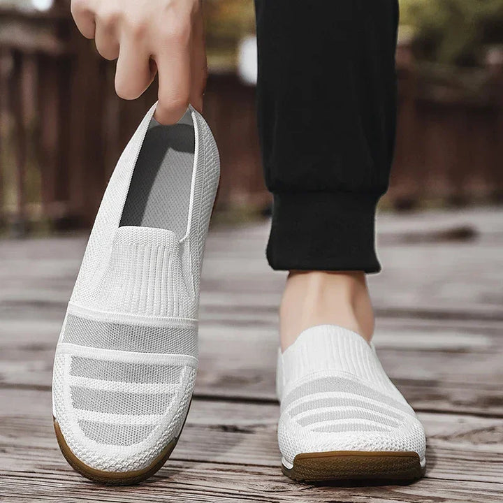 A person wearing black pants is putting on a white lightweight slip-on shoe with a brown sole while standing outdoors on a wooden surface. The other foot is already wearing a matching shoe.