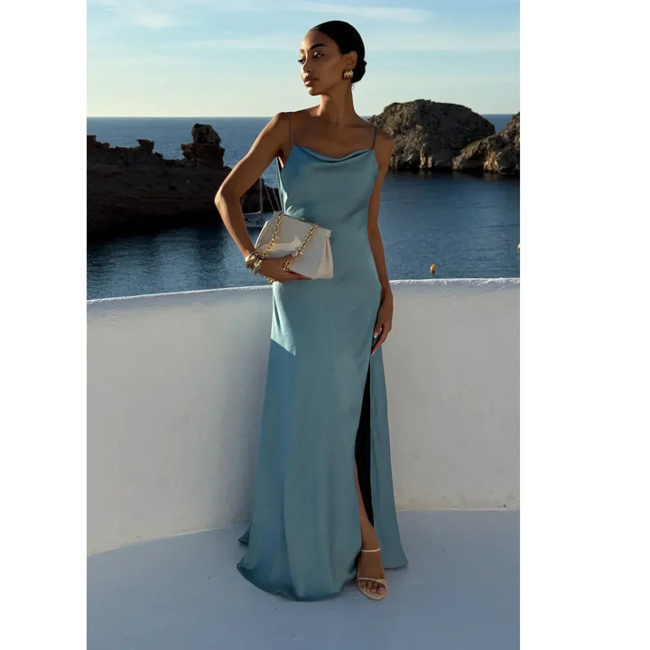 Elegant woman in a light blue satin gown by Rovelli, standing by the sea at sunset