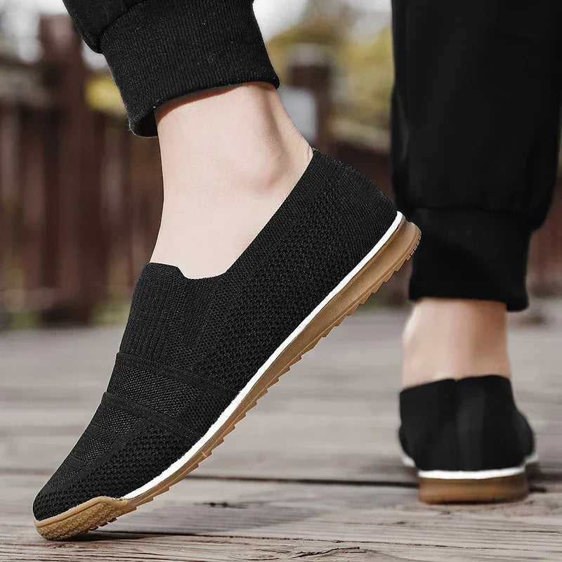 A person wearing lightweight slip-on shoes with tan rubber soles and black pants walks on a wooden outdoor surface.