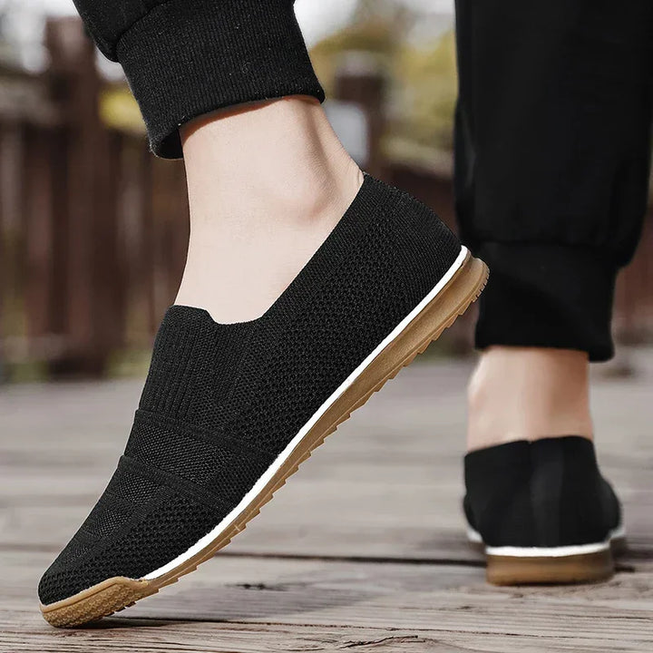 A person wearing lightweight slip-on shoes with tan rubber soles and black pants walks on a wooden outdoor surface.