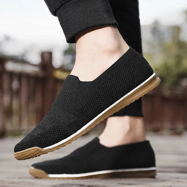 A person wearing lightweight slip-on knit shoes with white midsoles and brown rubber outsoles stands outdoors on a wooden surface, with trees and a fence blurred in the background.