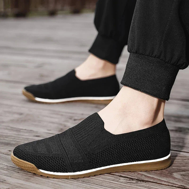 A person wearing black lightweight slip-on sneakers with white midsoles, brown outsoles, and a cushioned insole, paired with black jogger pants, standing on a wooden surface.