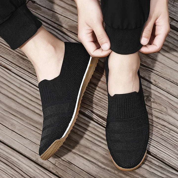 A person wearing lightweight slip-on knit sneakers with light brown soles and a cushioned insole is pulling up their pant leg while standing on a wooden floor.