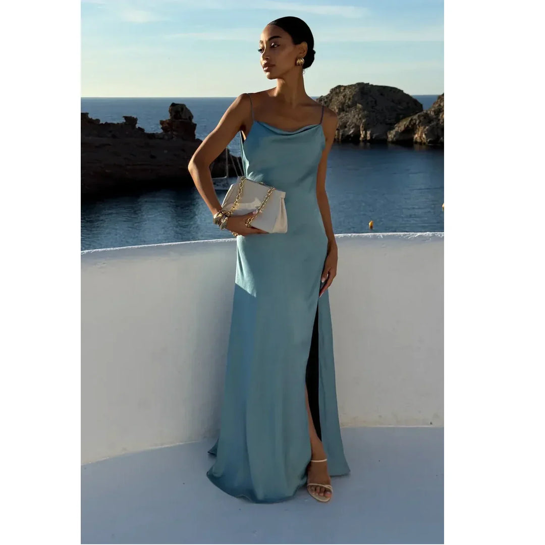 Elegant woman in a light blue satin dress holding a beige purse, standing by the sea at sunset