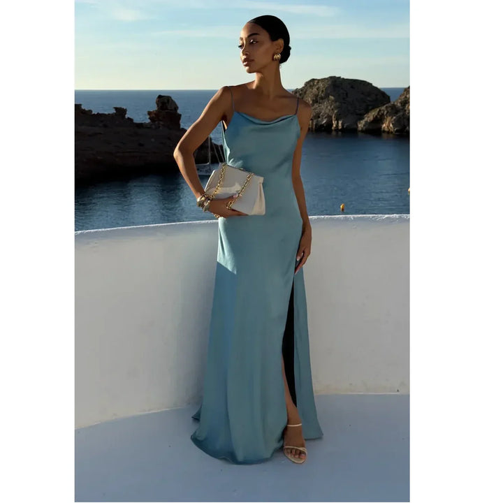 Elegant woman in a light blue satin dress holding a beige purse, standing by the sea at sunset