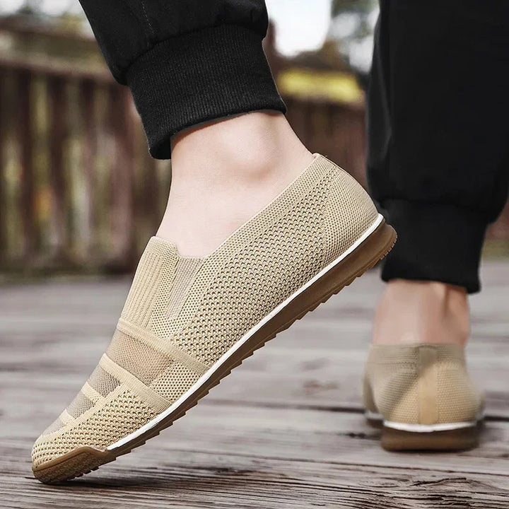 A person wearing lightweight slip-on knit shoes with a white sole is walking on a wooden surface outdoors, pants slightly rolled up at the ankles.