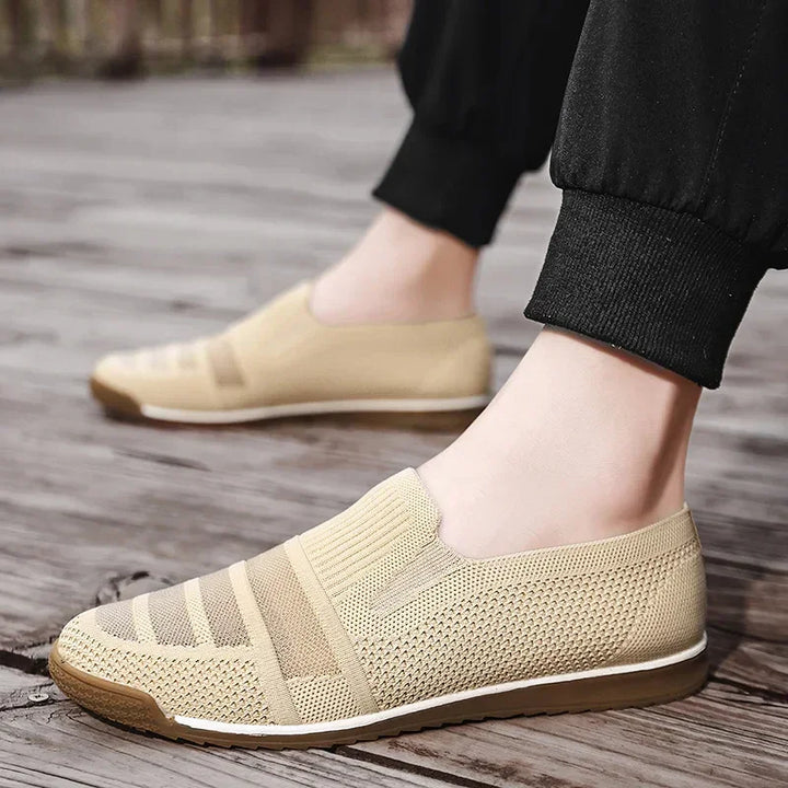 A person wearing lightweight slip-on mesh sneakers with cushioned insoles and brown soles stands on a wooden surface, their legs in black pants visible.