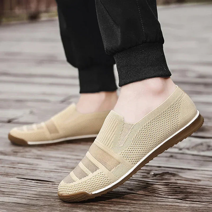 A person wearing lightweight slip-on mesh shoes with a textured design, cushioned insole, and brown soles stands on wooden planks outdoors. The person is dressed in black jogger pants.