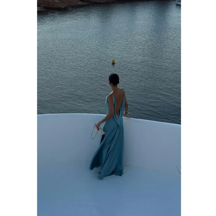 Woman in elegant blue dress with white purse overlooking water, Rovelli fashion.