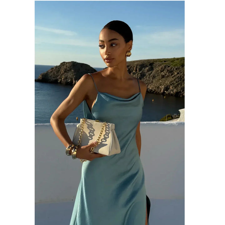 Woman in blue satin dress holding a gold chain bag, coastal background, Rovelli fashion