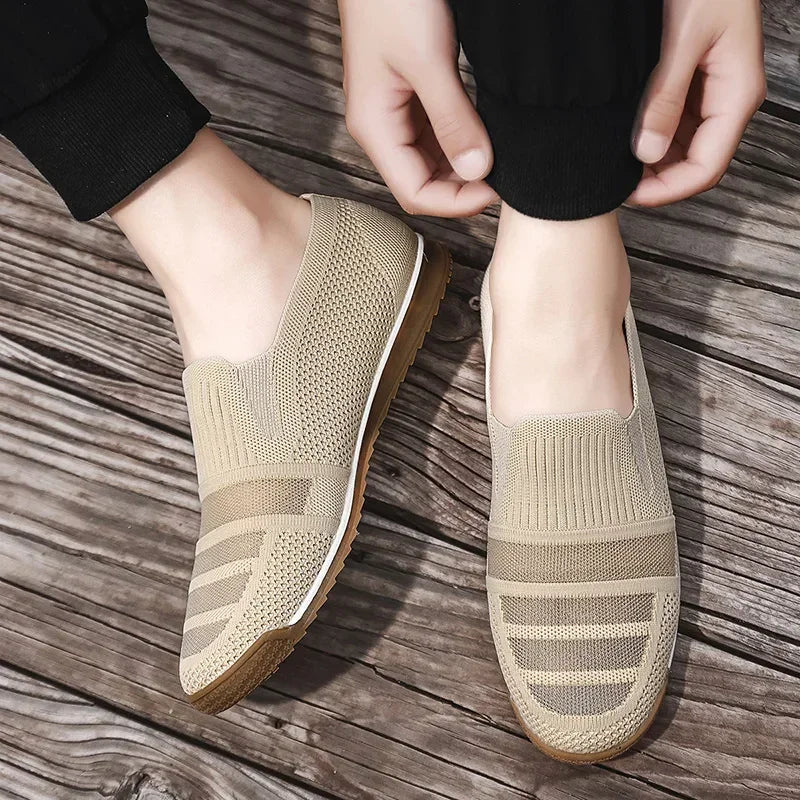A person wearing lightweight slip-on knit shoes with white soles stands on wooden flooring, adjusting the cuff of their black pants.