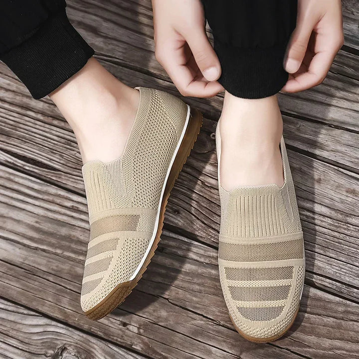 A person wearing lightweight slip-on knit shoes with white soles stands on wooden flooring, adjusting the cuff of their black pants.