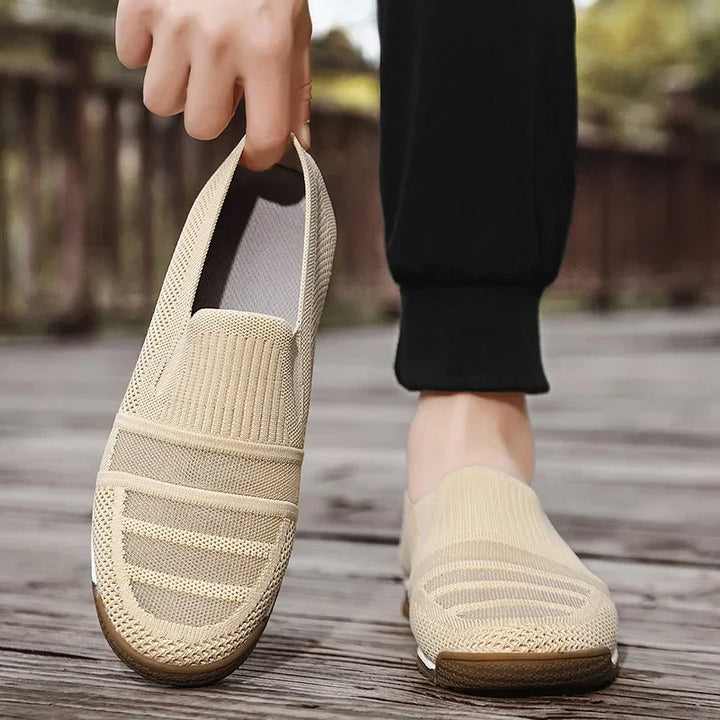 A person wearing lightweight slip-on shoes with a striped pattern, standing on a wooden surface outdoors, holding one shoe by the heel with their hand.